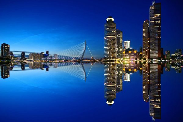 Beautiful night view of Rotterdam skyline with illuminated buildings and Erasmus Bridge reflection.